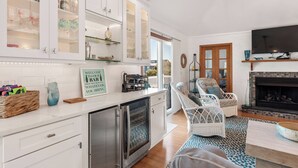 Une cuisine moderne avec des meubles blancs, un four encastré et un coin repas agrémenté de chaises en osier et d’une cheminée.