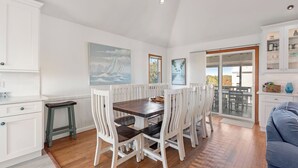 Un coin repas avec une table en bois et des chaises blanches, une cuisine blanche avec des placards, et une porte-fenêtre coulissante donnant sur une terrasse extérieure.