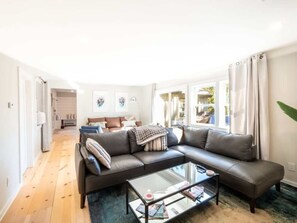 A modern living room with a sectional sofa, a glass coffee table, and a view of the outdoors through large windows. - Renovated Downtown Great Barrington House (Great Barrington)