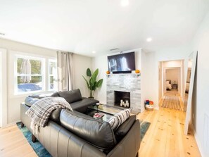 A modern living room with a leather sofa, a glass coffee table, a fireplace, and a flat-screen TV mounted above it. - Renovated Downtown Great Barrington House (Great Barrington)