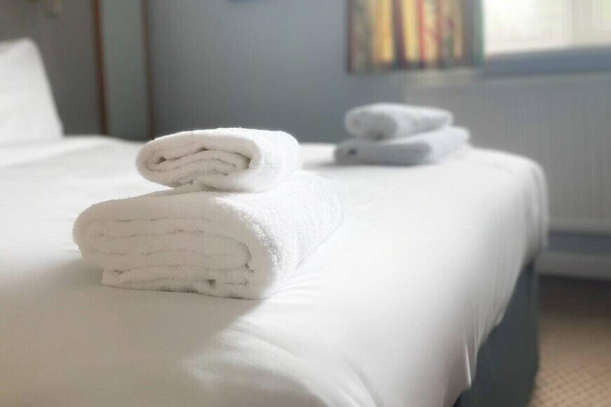 A neatly made bed with folded white towels, a window with striped curtains, and a framed picture on the wall.
