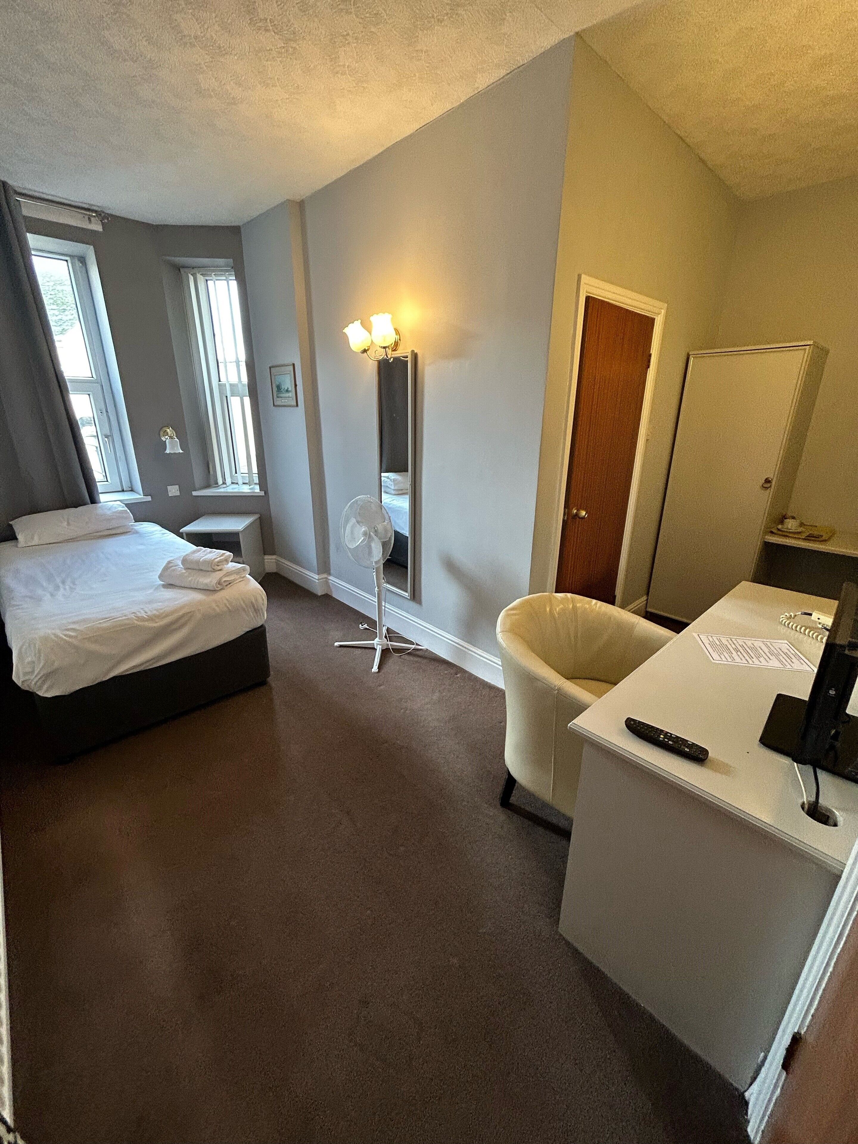 Quarto de hotel com uma cama, uma escrivaninha, uma cadeira, luminária de mesa, ventilador e um guarda-roupa.