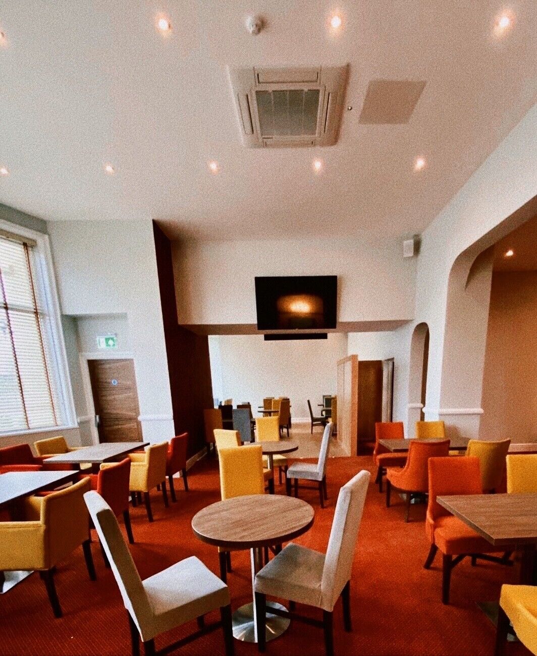Un grand espace dédié à la salle à manger, avec plusieurs tables et chaises, un téléviseur à écran plat fixé au mur et une bouche d’aération au plafond.