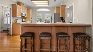Uma cozinha ampla com armários de madeira, uma ilha grande com banquetas, iluminação pendente e vista pela janela.