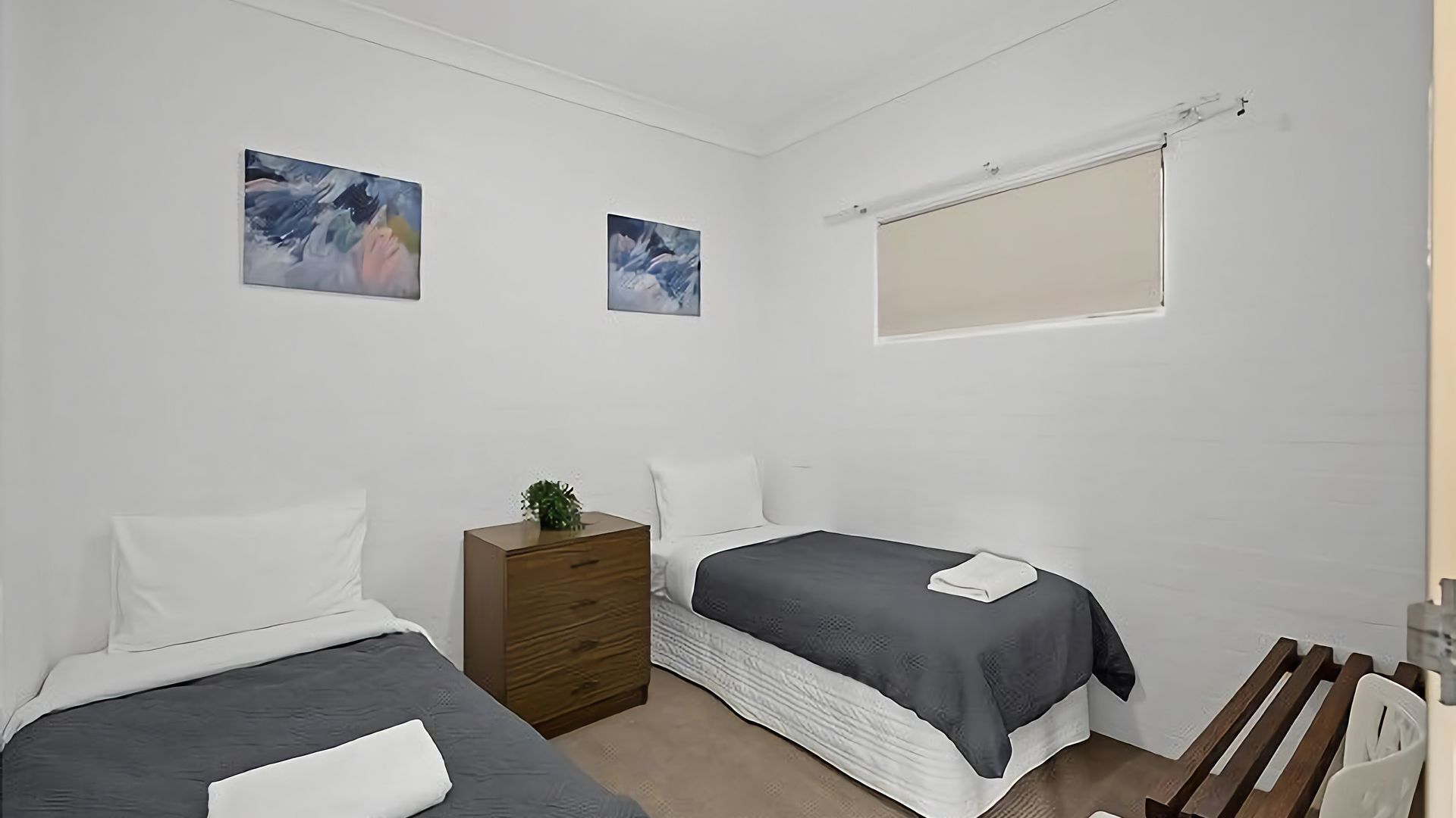 A small, clean room with two beds, a wooden dresser, and two framed pictures on the wall.
