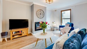 A modern living room with a flat-screen TV, a blue sofa, a small round table with a vase of flowers, and a large window. - 2 Bed in Braunton (oc-stepco) (Braunton)