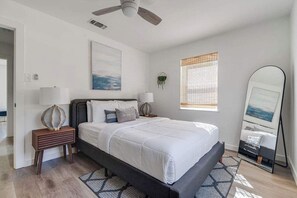 Een slaapkamer met een bed, nachtkastjes, een plafondventilator, een raam met jaloezieën, een spiegel aan de muur en een schilderij.