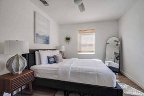 A modern bedroom with a large bed, bedside tables, a wall-mounted mirror, and a window with blinds.