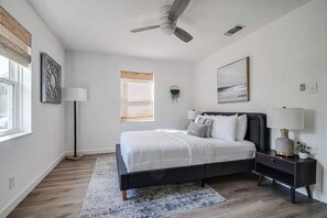 Un dormitorio con cama, mesitas de noche, ventilador de techo, ventana con persianas y una obra de arte en la pared.