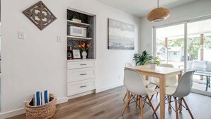A modern kitchen with white cabinetry, a dining table with chairs, a hanging light fixture, and a view through a large window. - Charming Getaway 4 Min to Downtown-Hot Tub, Firepit Grill (Fredericksburg)