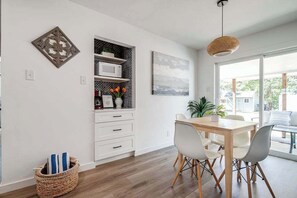 A modern kitchen with white cabinetry, a dining table with chairs, a hanging light fixture, and a view through a large window.