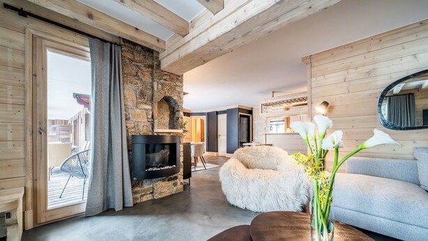 Una sala moderna con chimenea de piedra, un sofá, un otoman suave y un jarrón con flores.