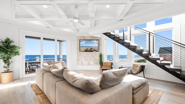 Ein geräumiges Wohnzimmer mit einem großen, beigefarbenen Ecksofa, einer Treppe, die zu einer oberen Ebene führt, und Blick aufs Meer durch große Fenster.