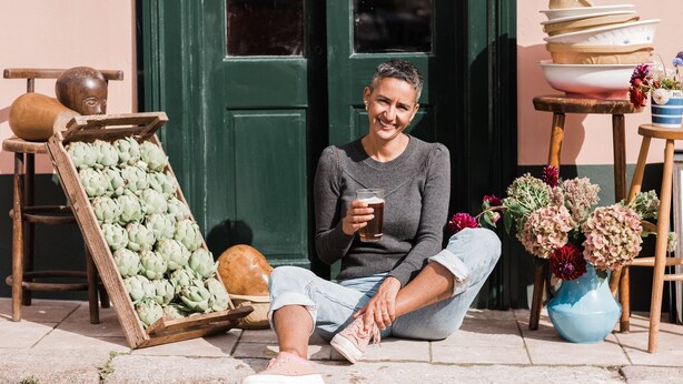 A person sitting on the ground outside a building with a green door, holding a glass of beer. Artichokes are displayed in a wooden crate, and there are flowers in a vase. - The Bull Inn (Totnes)
