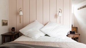 A neatly made bed with white linens and two pillows, a bedside table with a lamp, a small potted plant, and a wooden nightstand with a clock. - The Bull Inn (Totnes)