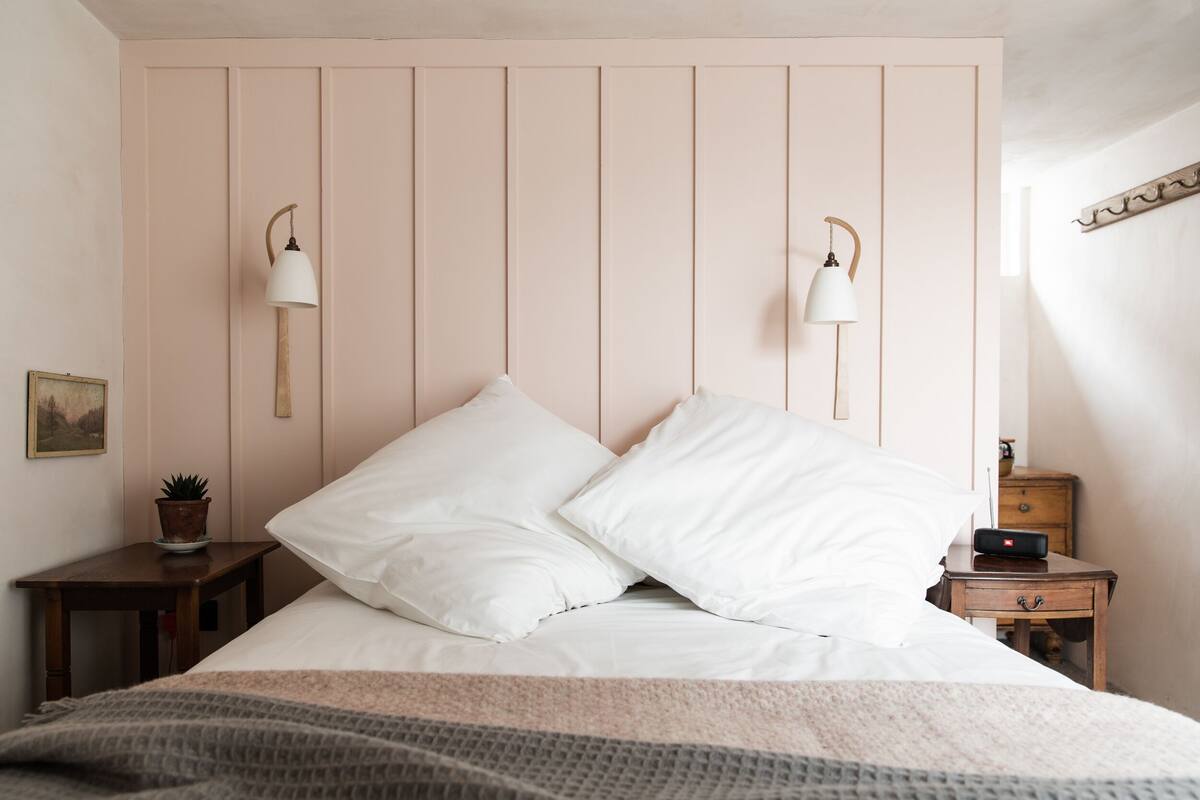 A neatly made bed with white linens and two pillows, a bedside table with a lamp, a small potted plant, and a wooden nightstand with a clock.