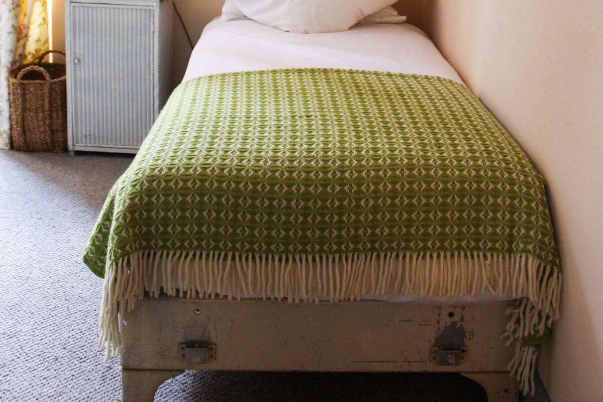 A small, neatly made bed with a green patterned blanket, a white pillow, and a bedside table with a lamp and a small basket.