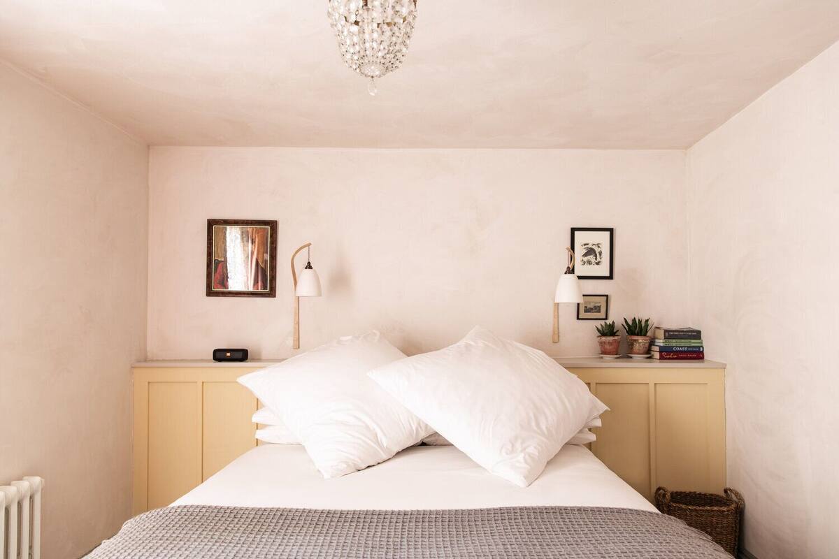 A bedroom with a bed, bedside lamps, a nightstand, and a small table with books and plants.