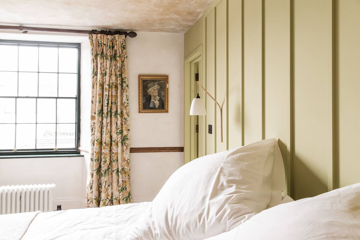 A bedroom with a bed, a window with floral curtains, a radiator, and a picture on the wall.