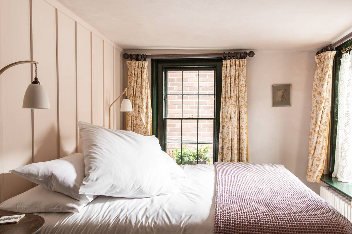 A neatly made bed with white linens and a patterned throw blanket, a bedside lamp, and a window with floral curtains.
