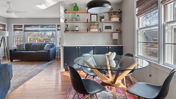 A modern living room with a glass dining table, black chairs, a blue sofa, and a kitchen area with wooden shelves and a hanging light. - Walk to Parks- Pet Friendly-Top Neighborhood (Rapid City)