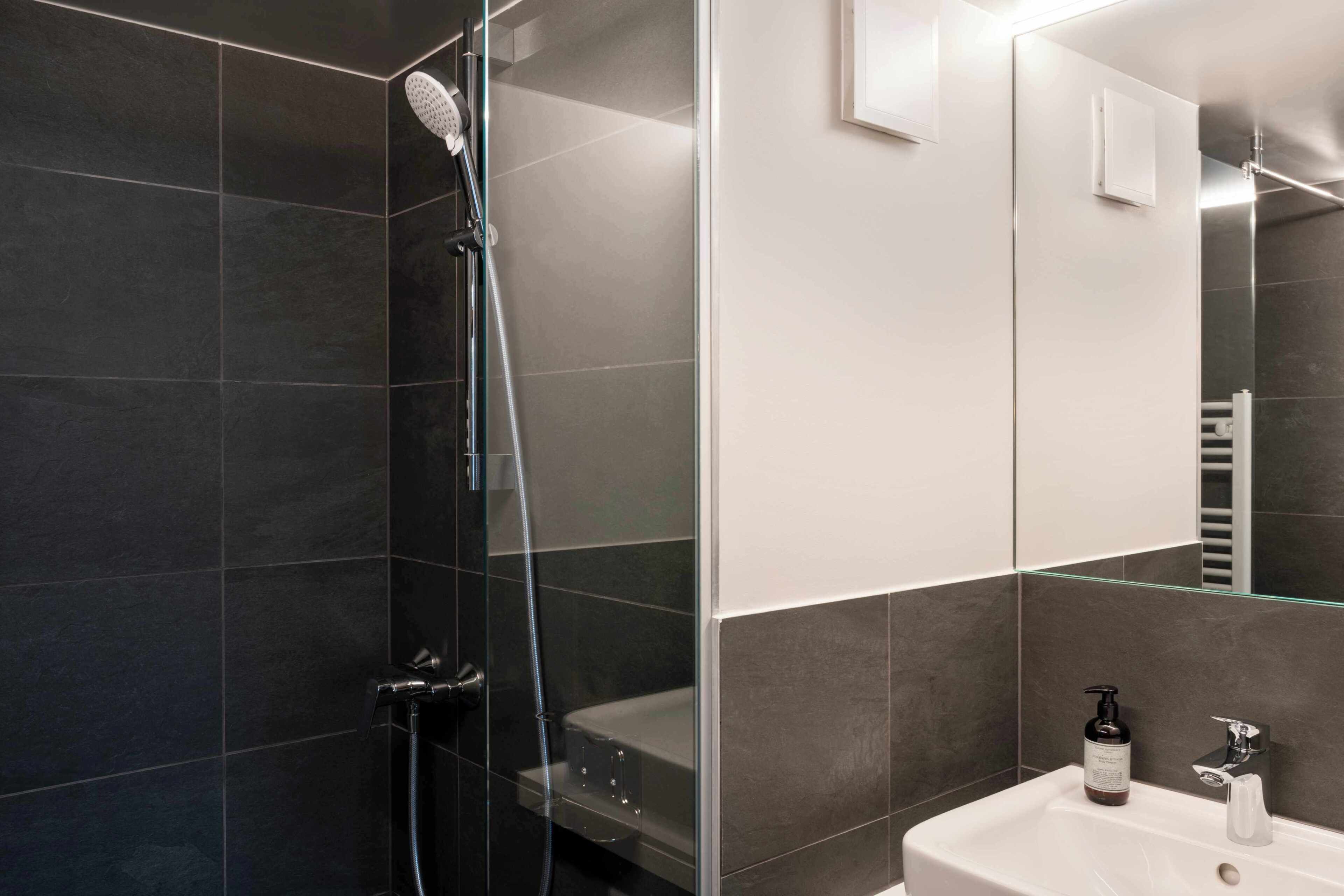 A modern bathroom with dark tiles, a glass shower enclosure, a white sink, and a mirror.