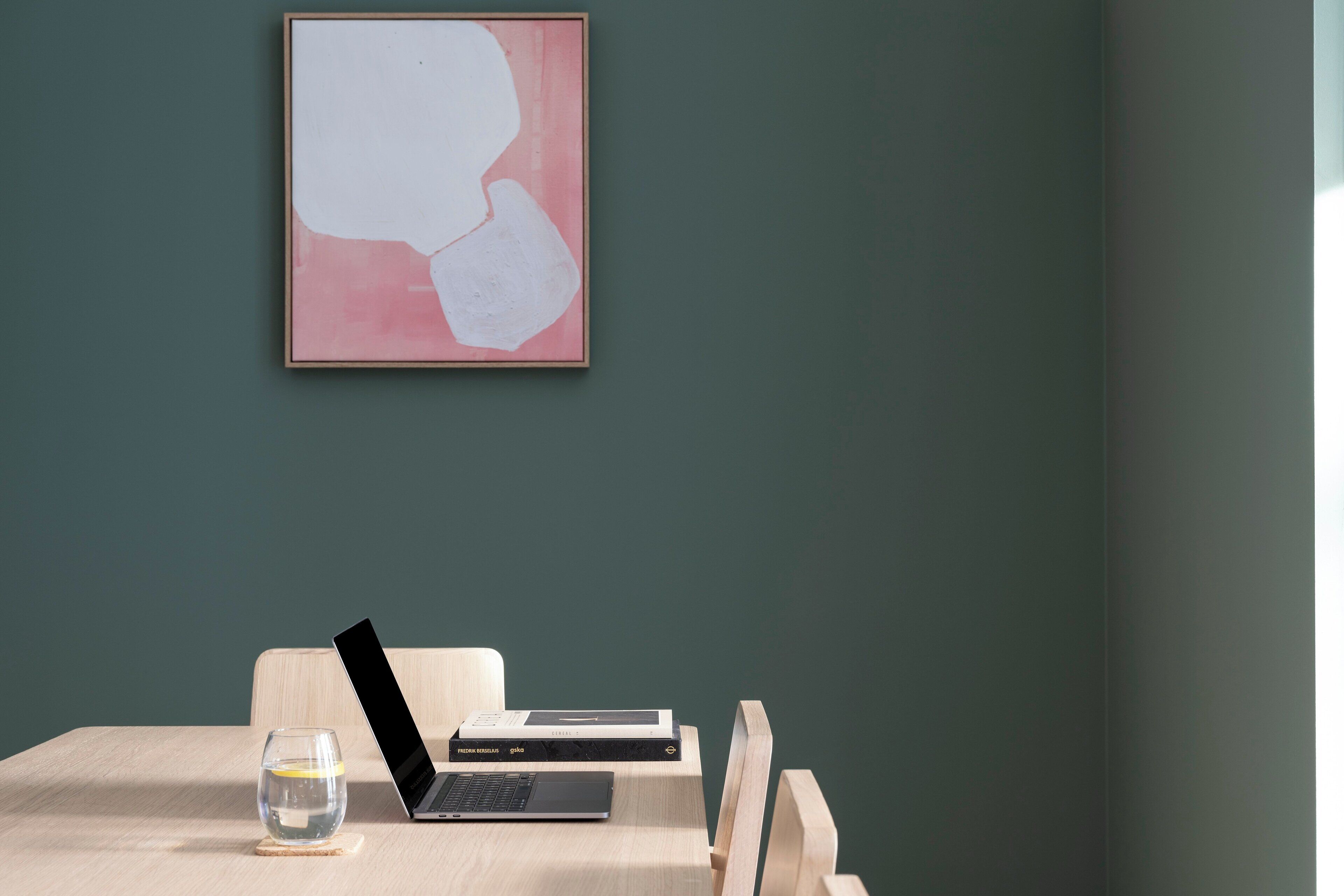 Une pièce au décor minimaliste, avec un ordinateur portable posé sur une table en bois, un verre d’eau et un tableau abstrait encadré accroché au mur.