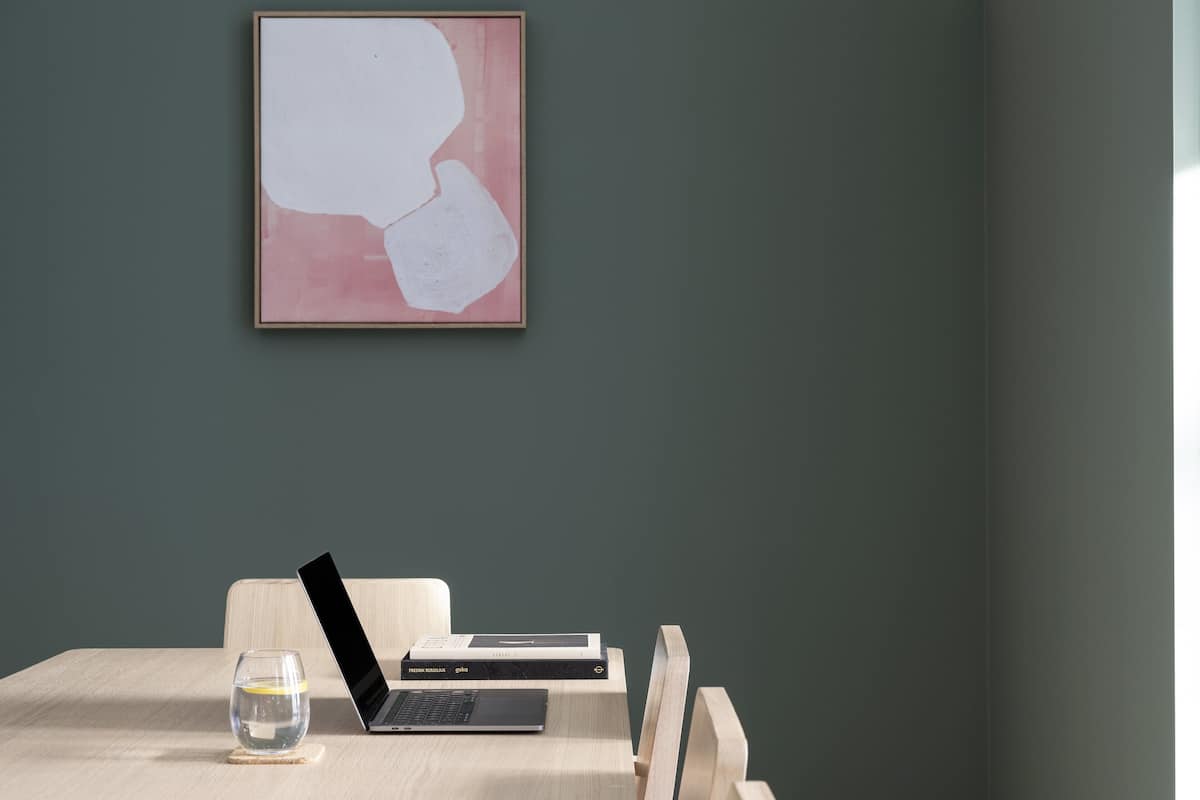 Une pièce au décor minimaliste, avec un ordinateur portable posé sur une table en bois, un verre d’eau et un tableau abstrait encadré accroché au mur.