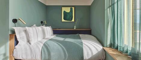 A bedroom with a bed, bedside lamps, a window with curtains, a wall-mounted artwork, and a small table with a vase.