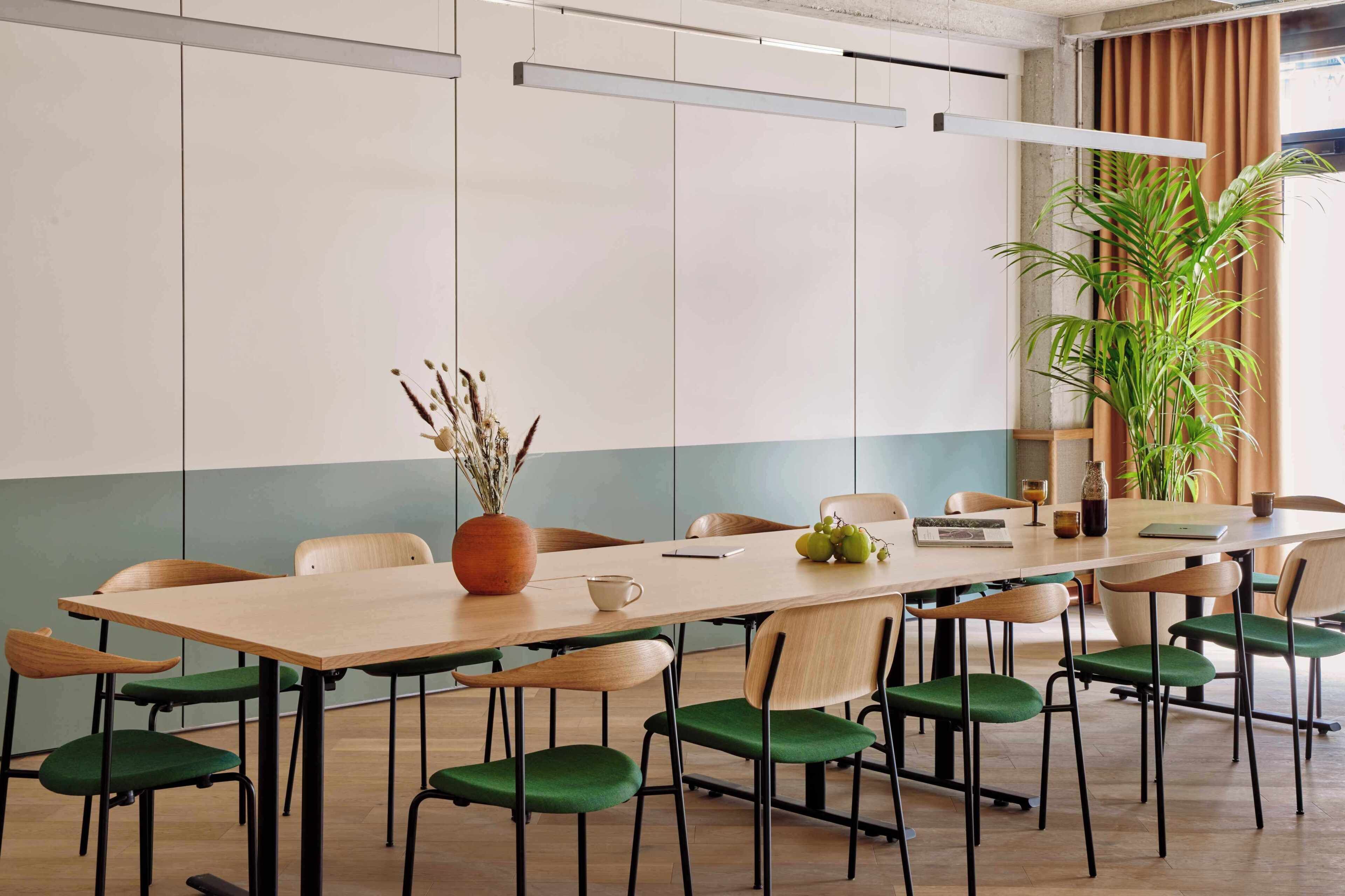 A conference room with a long table, green chairs, a potted plant, and a window with curtains.