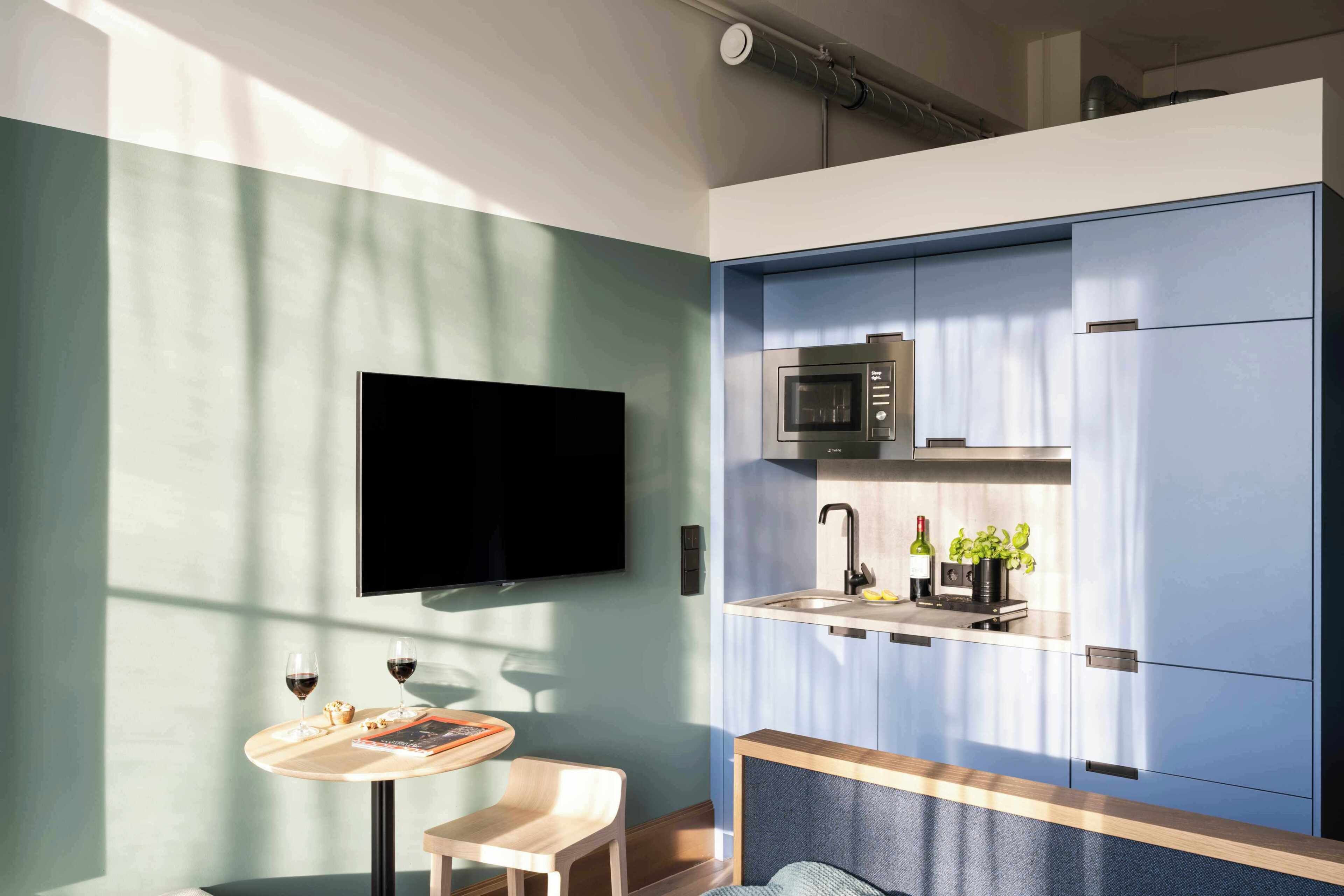 A compact kitchen with a microwave, sink, and wine bottle. There's a flat-screen TV on the wall and a small table with a chair.