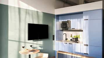 A compact kitchen with a microwave, sink, and wine bottle. There's a flat-screen TV on the wall and a small table with a chair.
