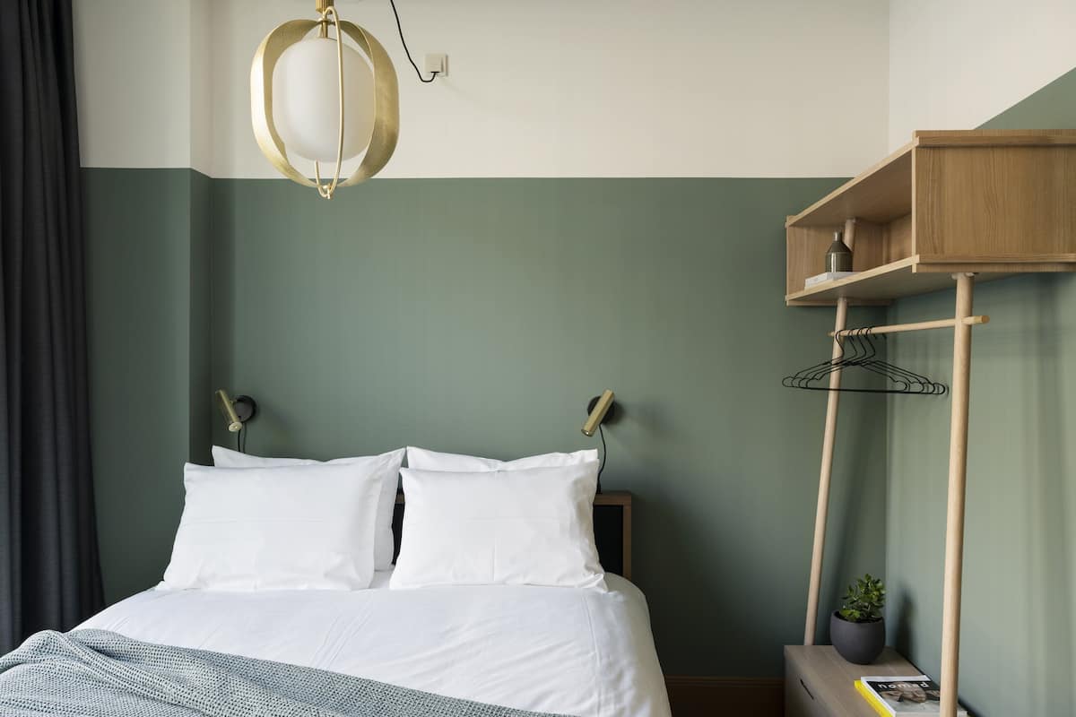 Un lit bien fait, avec du linge de lit blanc, un luminaire suspendu, une étagère en bois ornée d’une plante et un magazine posé sur la table de chevet.