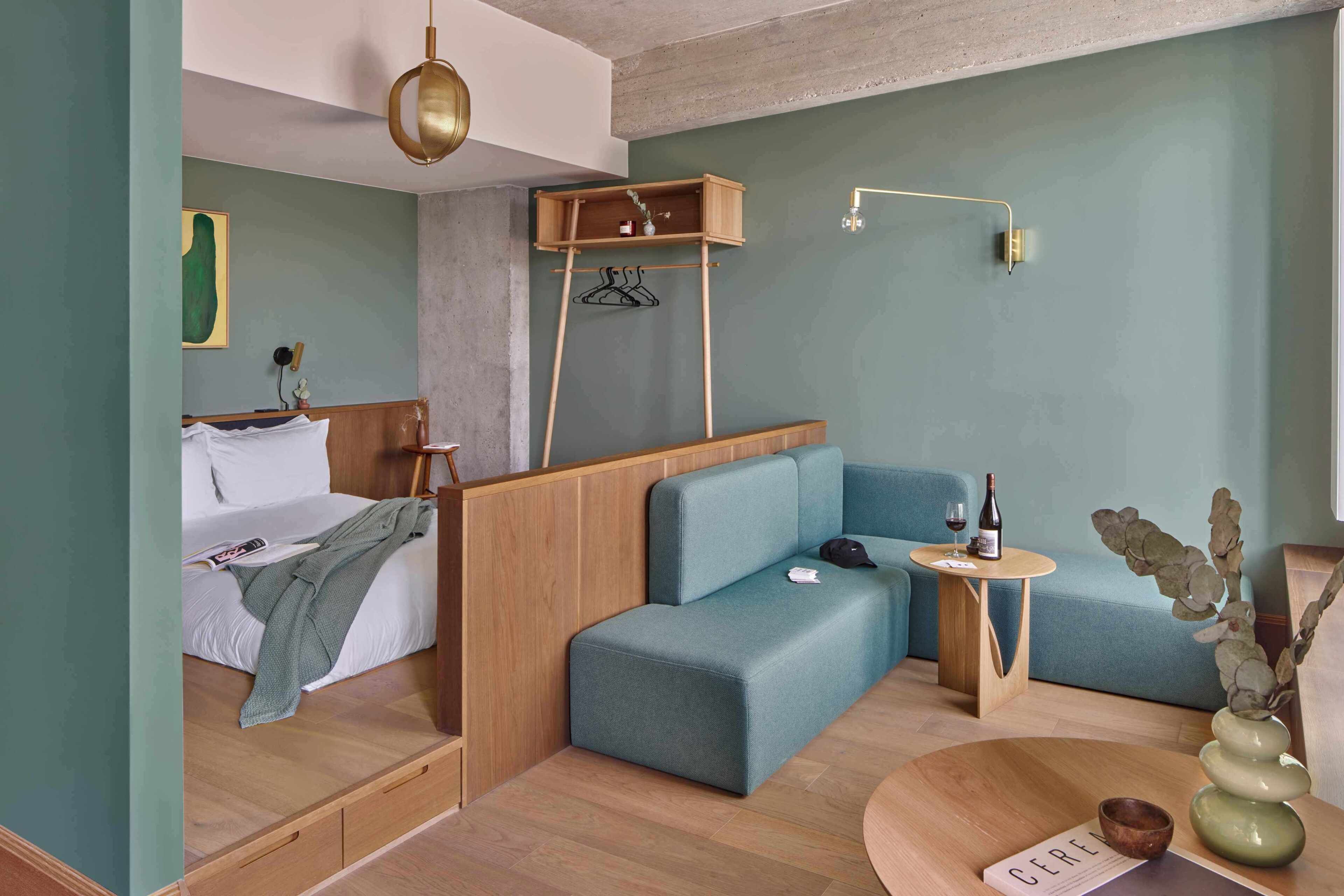 Habitación de hotel pequeña con cama, sofá azul, una mesita y un estante de madera con perchero.