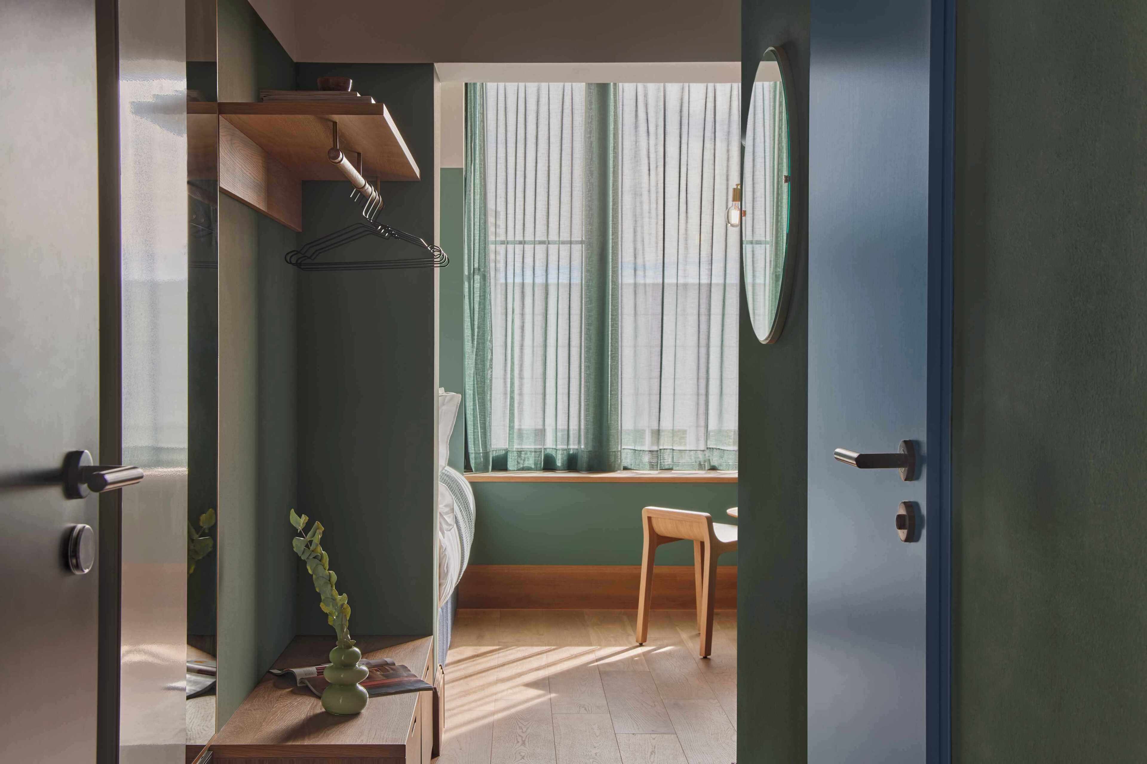 Ein Zimmer mit einem Bett, einem Sessel, einem Fenster mit Vorhängen, einem Kleiderhaken und einem kleinen Tisch mit einer Pflanze.