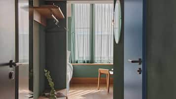 Ein Zimmer mit einem Bett, einem Sessel, einem Fenster mit Vorhängen, einem Kleiderhaken und einem kleinen Tisch mit einer Pflanze.