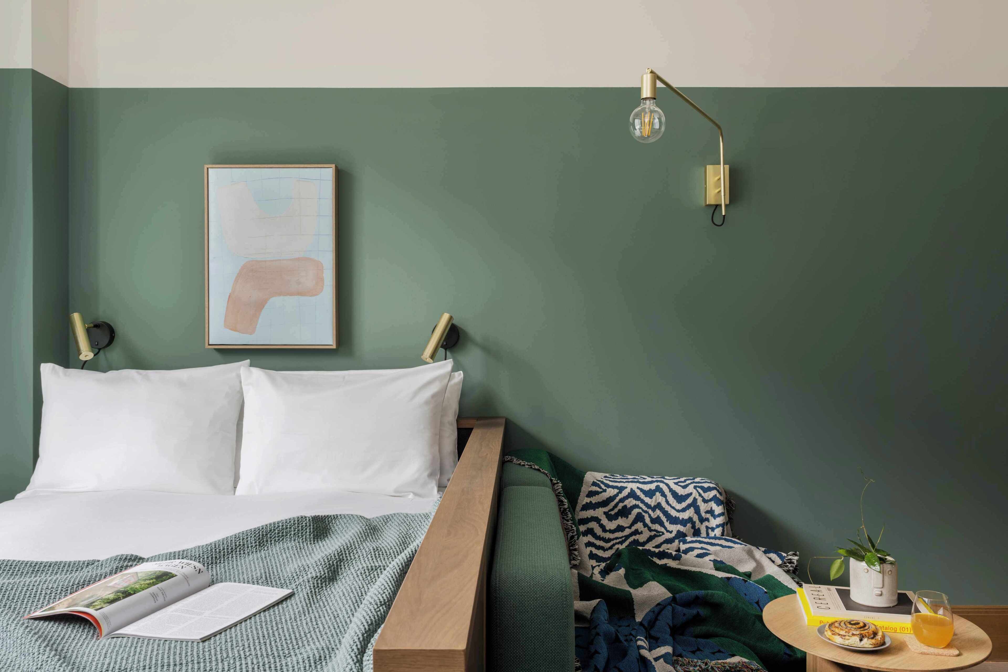 A neatly made bed with a magazine, a bedside lamp, and a small table with a plant and a glass of juice.