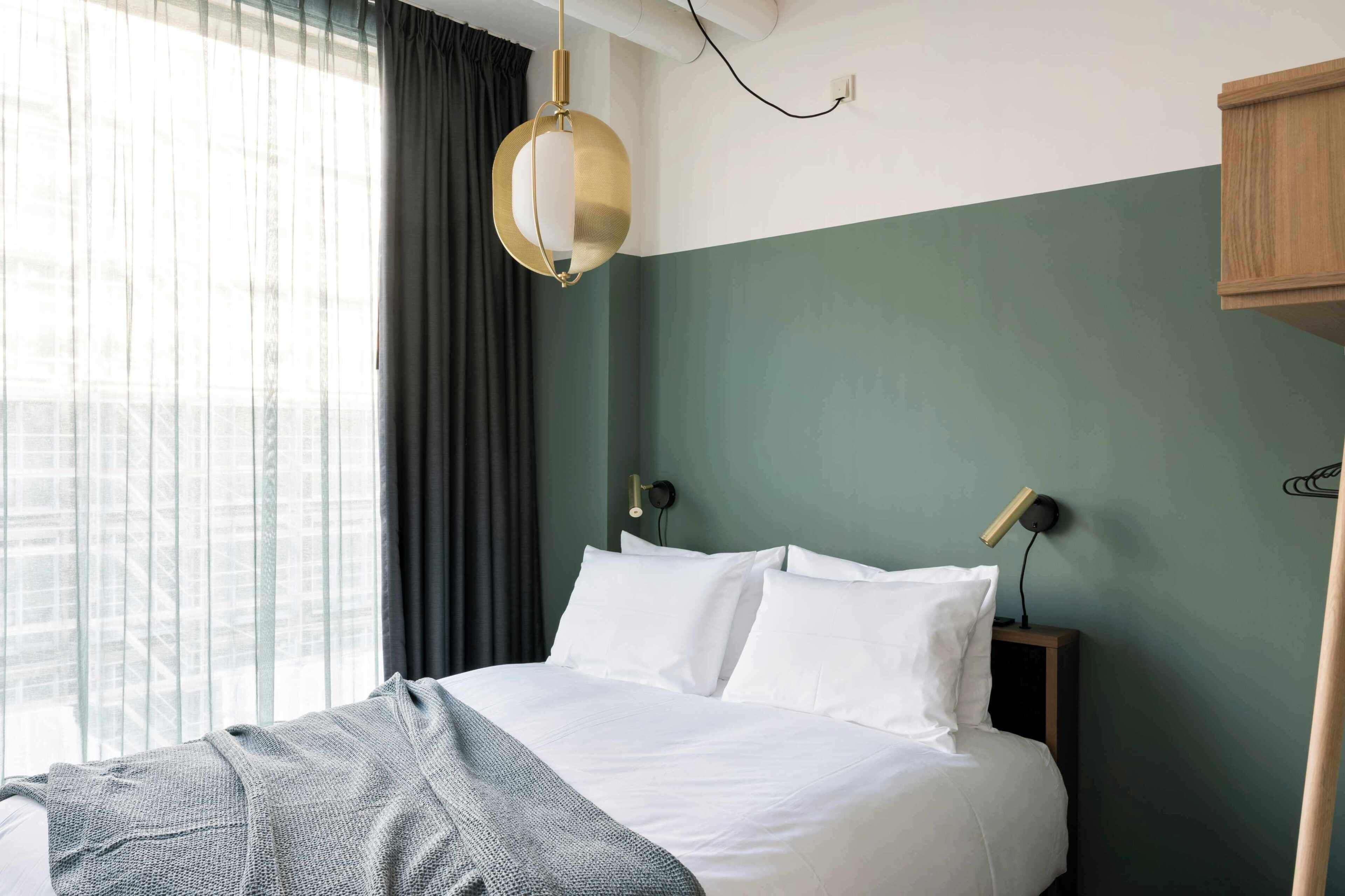 A hotel room with a bed, a window with curtains, a wall-mounted lamp, and a wooden cabinet.