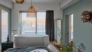 A modern hotel room with a large bed, a bathtub, a hanging light fixture, and a view of the city. - WunderLocke Munich (Munich)