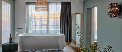 A modern hotel room with a large bed, a bathtub, a hanging light fixture, and a view of the city.