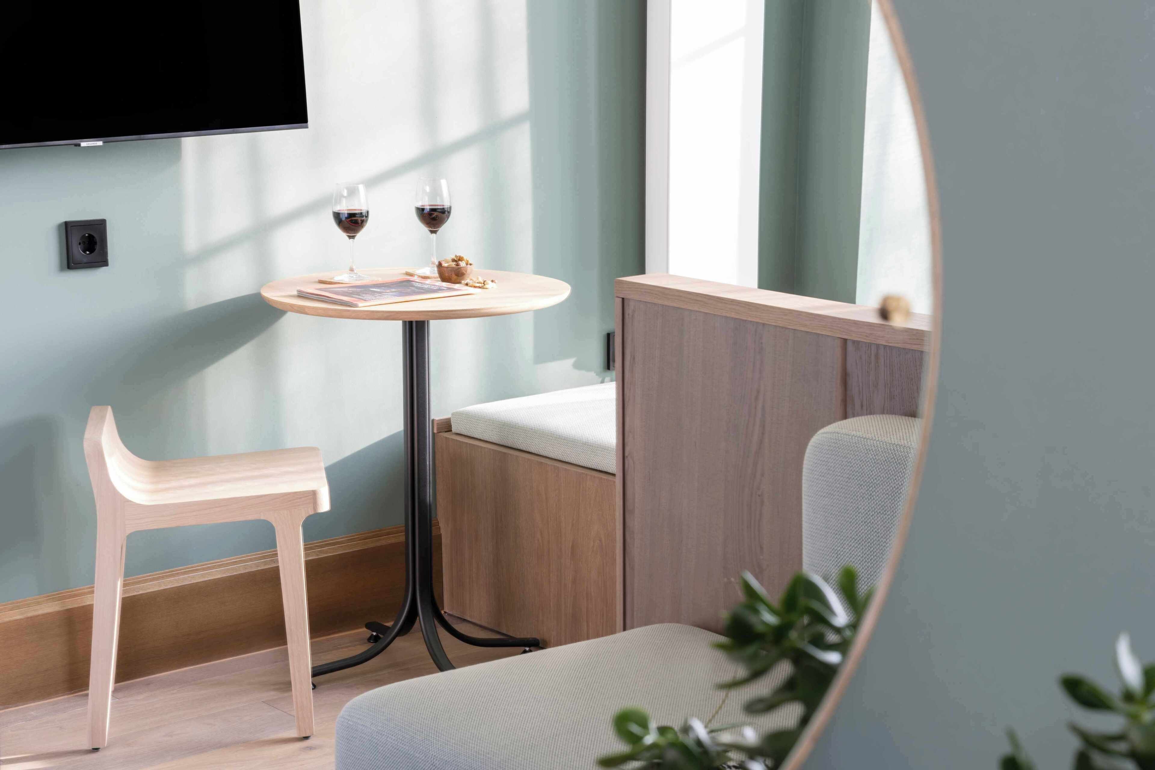 Una habitación moderna con una silla de madera, una mesita redonda con dos copas de vino, un sofá y un televisor montado en la pared.