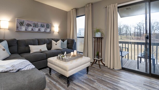 Un salon avec un canapé d’angle gris, un pouf blanc, un tableau représentant des plantes succulentes, une lampe sur pied et une vue sur l’extérieur grâce à une porte-fenêtre coulissante.