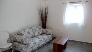 Ein Wohnzimmer mit einem gemusterten Sofa, einem Holztisch, einem weißen Sessel und einer hohen Zierpflanze.