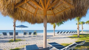 A beach with a thatched umbrella, lounge chairs, and palm trees. - Pointe Santo C6 | Upgraded Walk Out Beach Residence | Fantastic Amenities and Gulf Views (Sanibel)