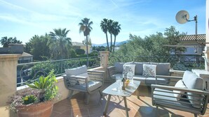 A terrace with outdoor furniture, a potted plant, and a view of palm trees and mountains. - Oliveres - Six Bedroom Villa, Sleeps 12 (Pollença)