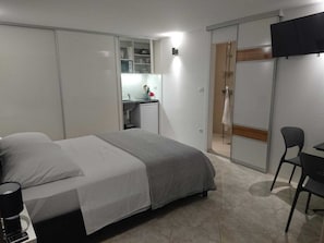 A bedroom with a bed, a desk with a chair, a wardrobe, and a shower area.
