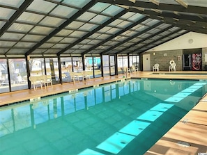 An indoor swimming pool with clear glass walls, wooden deck, and white lounge chairs.