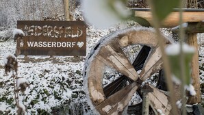 Un cartello di legno rustico con il testo "Wasserdorf" e un simbolo a forma di cuore, sullo sfondo di un paesaggio innevato con una grande struttura circolare in legno in primo piano.