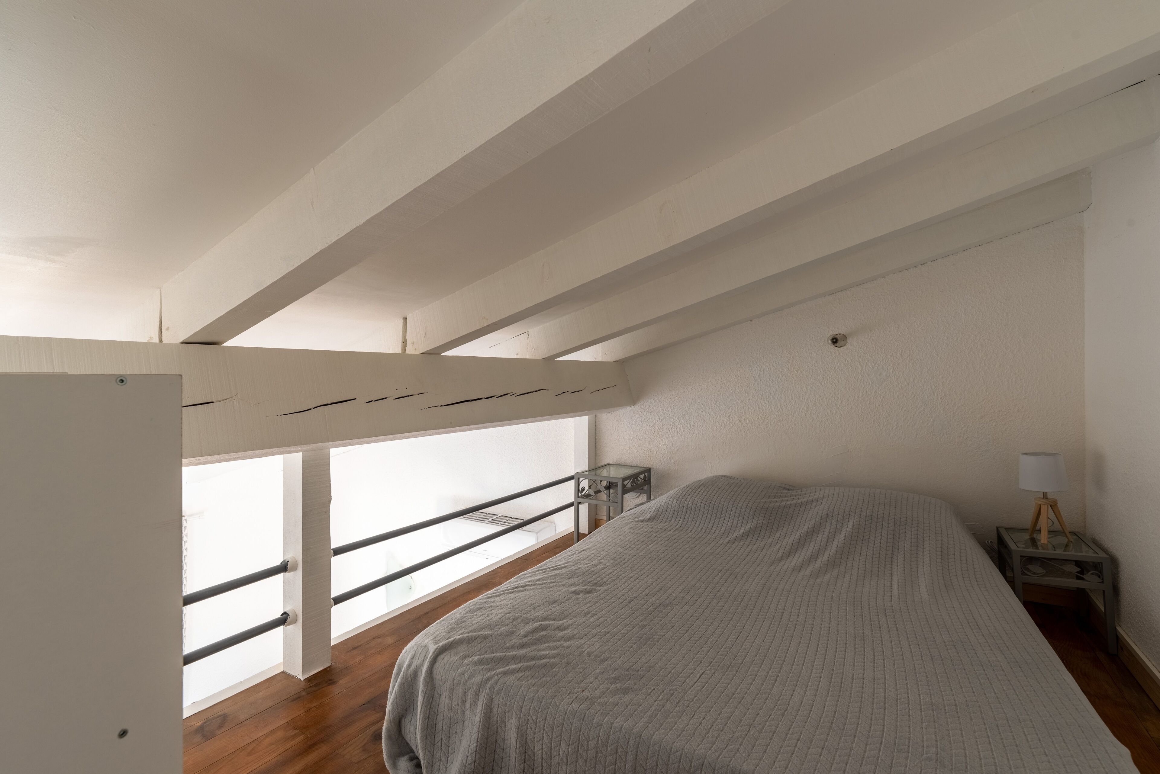 A bedroom with a bed, a nightstand, a window with a view, and a white ceiling with beams.
