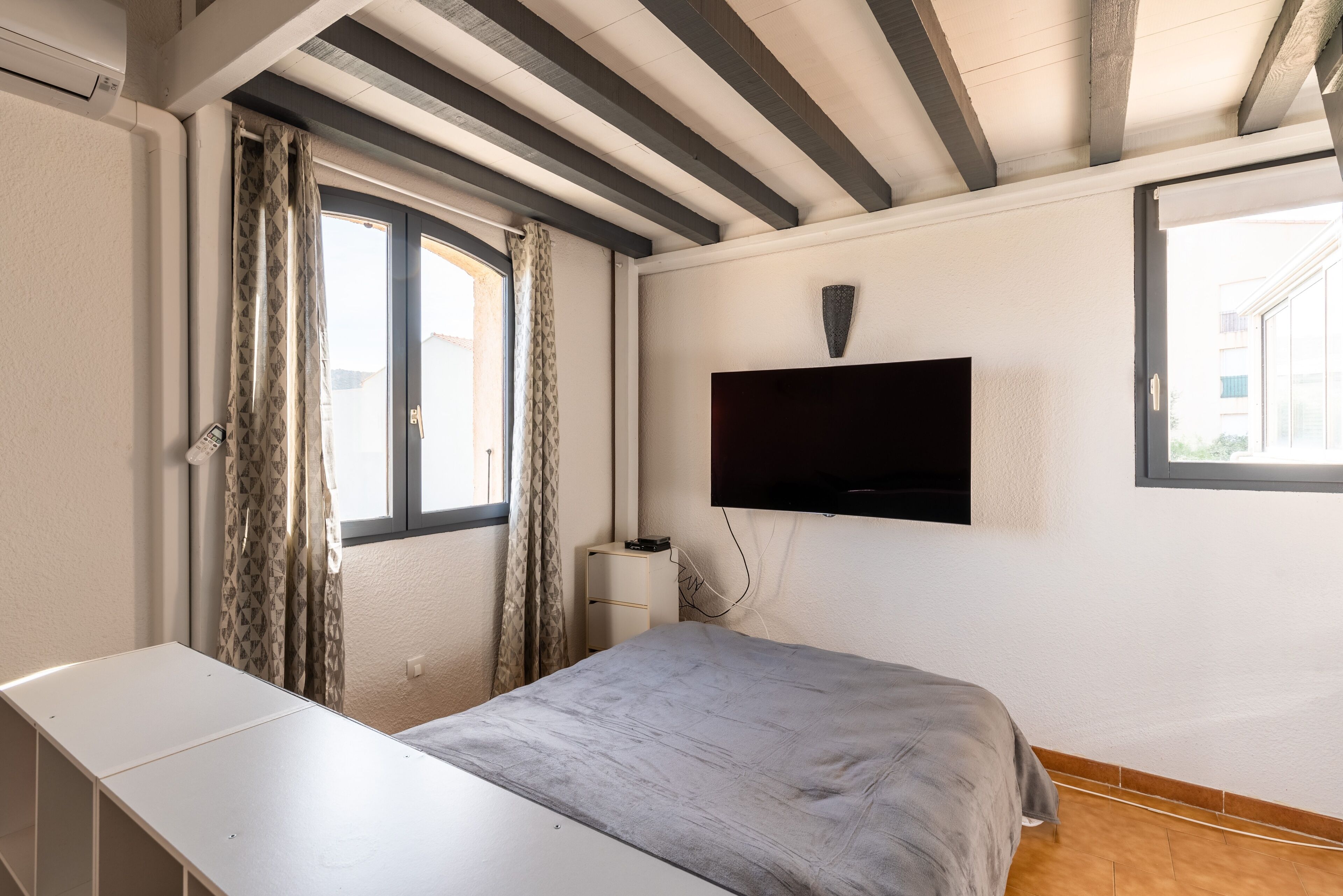 Un dormitorio con cama, televisión montada en la pared, ventana con cortinas y un escritorio blanco.